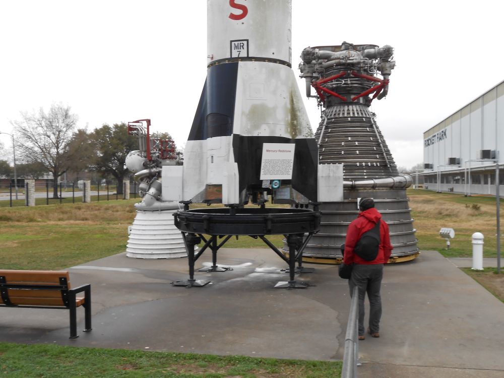 rocket park at johnson space center on a cloudy day. the bottom of a Mercury-Redstone rocket (which is about as wide as an adult’s outstretched arms) is sitting between two massive rocket engines: the smaller of them (the upper stage engine of the Saturn V, the J-2) is on the left, and it’s wider than the entire Redstone. The larger engine (the first stage engine of the Saturn V, the F-1) is twice as large as the J-2. An adult is standing looking at all three, and it appears that five people their size could fit inside the engine bell of F-1