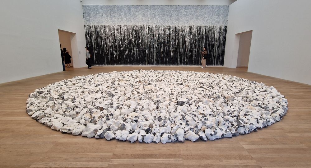 A large filled circle of white and grey flint on the floor of a gallery, with a painting filling the wall behind it. The painting is black for the lower two thirds, while the upper third is whitewashed and has visible hand swirls in the wash. The wash has dripped along the width of the paiting, causing white streaks into the back area below it. 