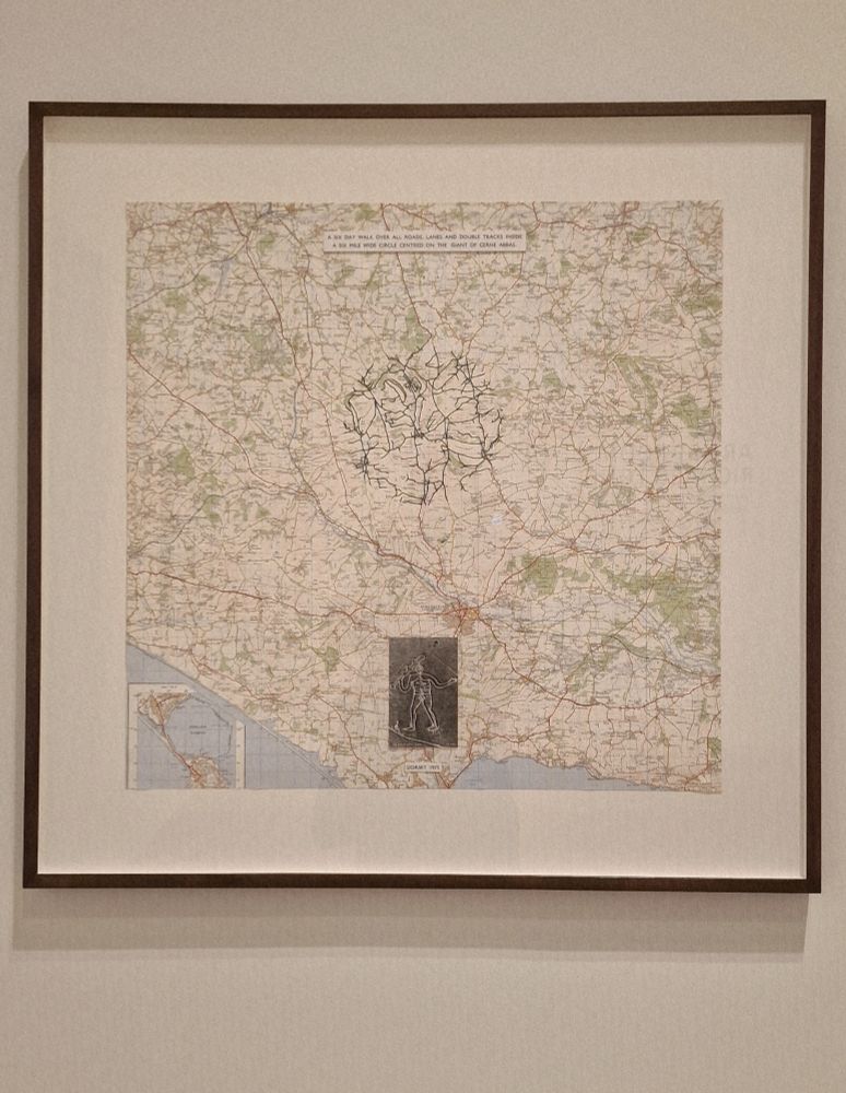 A framed UK Ordinance Survey map, centred on Cerne Abbas. A circular area in the centre of the map has all of the roads and tracks filled in with black ink. Below the black design is a photo of the Cerne Abbas giant, a figure approximately 55 metres tall, cut into turf and filled with chalk. Above the design is a printed label stating "A six day walk, over all roads, lanes and double tracks inside a six mile wide circle centred on the giant of Cerne Abbas." 