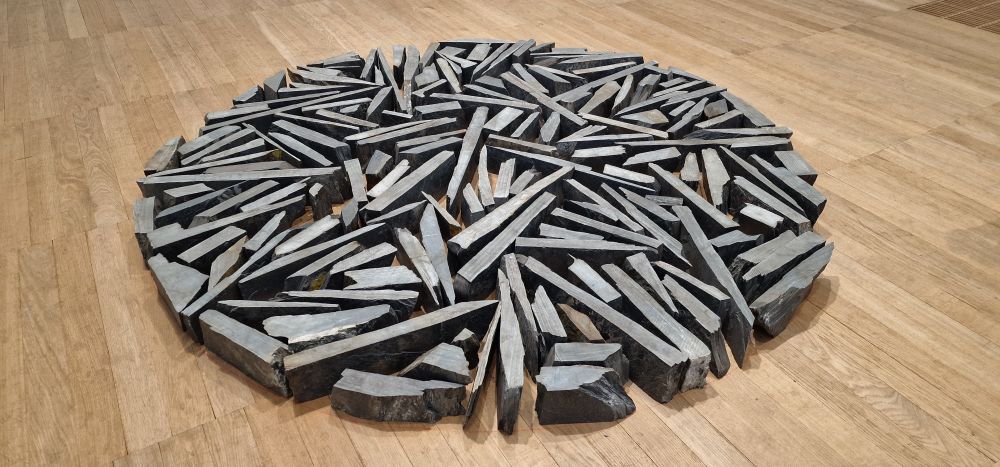 Multiple lengths of slate pointing in different directions but within a circular boundary on the floor of a gallery. 