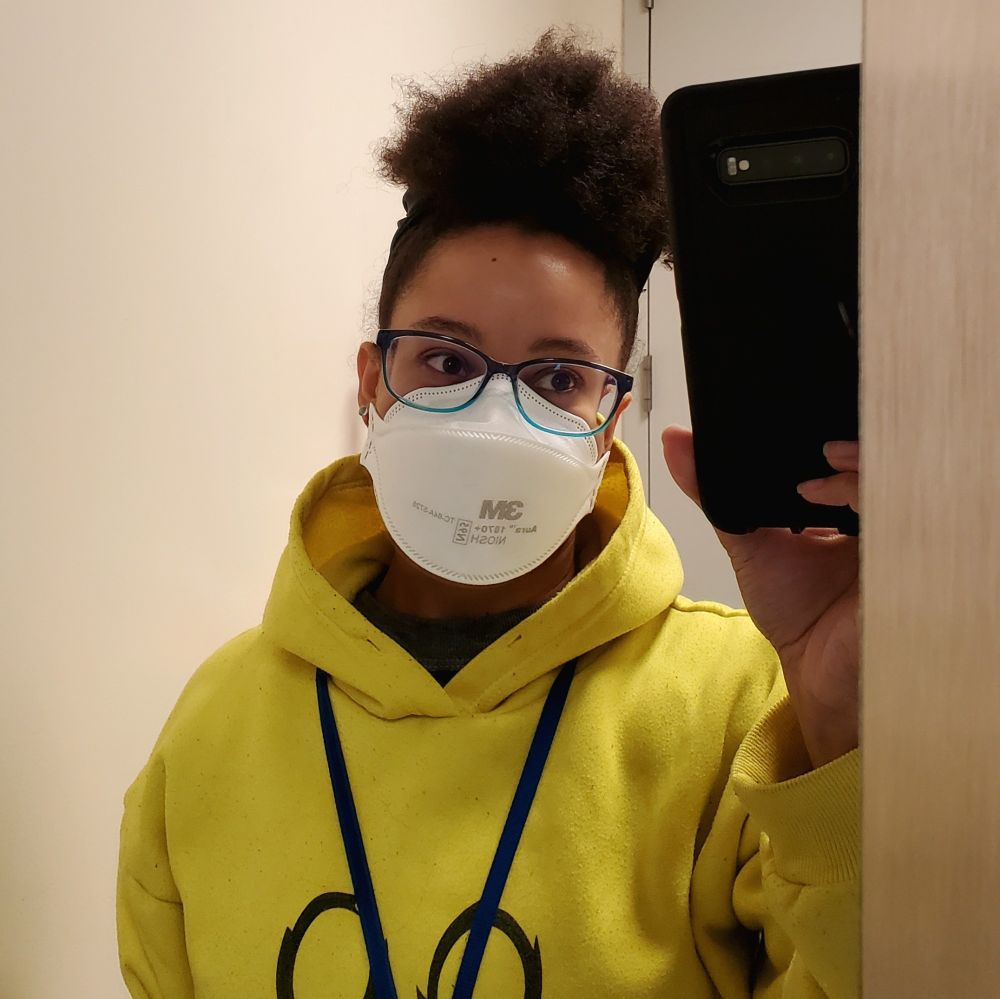 Bathroom selfie wearing yellow hoodie, blue glasses, KN95 mask, hair in puff ball on top of head.
