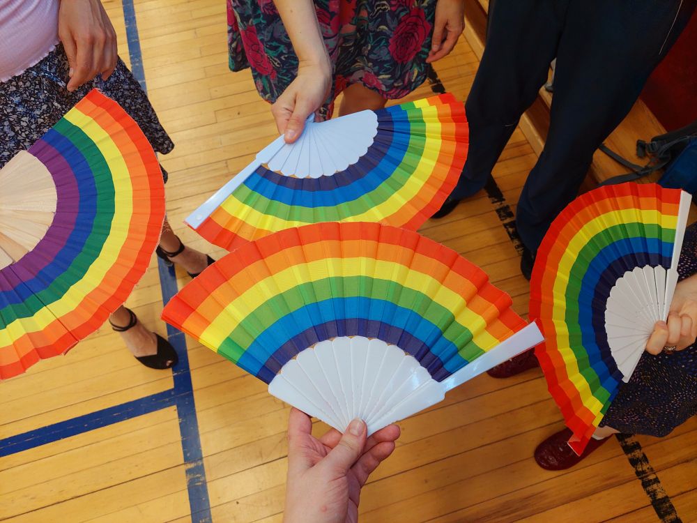 Photo of four fans in rainbow colors held by people wearing dance shoes. The wooden floor has the markings of a school gym.