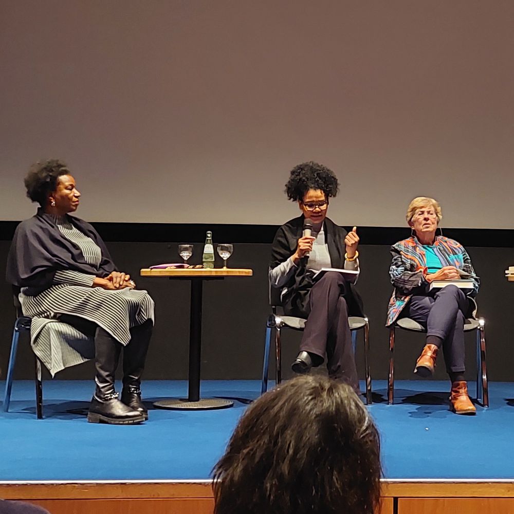 Photo of the Black activists Abeena Ademako (left) & Glenda Obermuller (middle) together on stage with the queer filmmaker Dagmar Schulz (right). Glenda Obermuller speaks into a microphone.