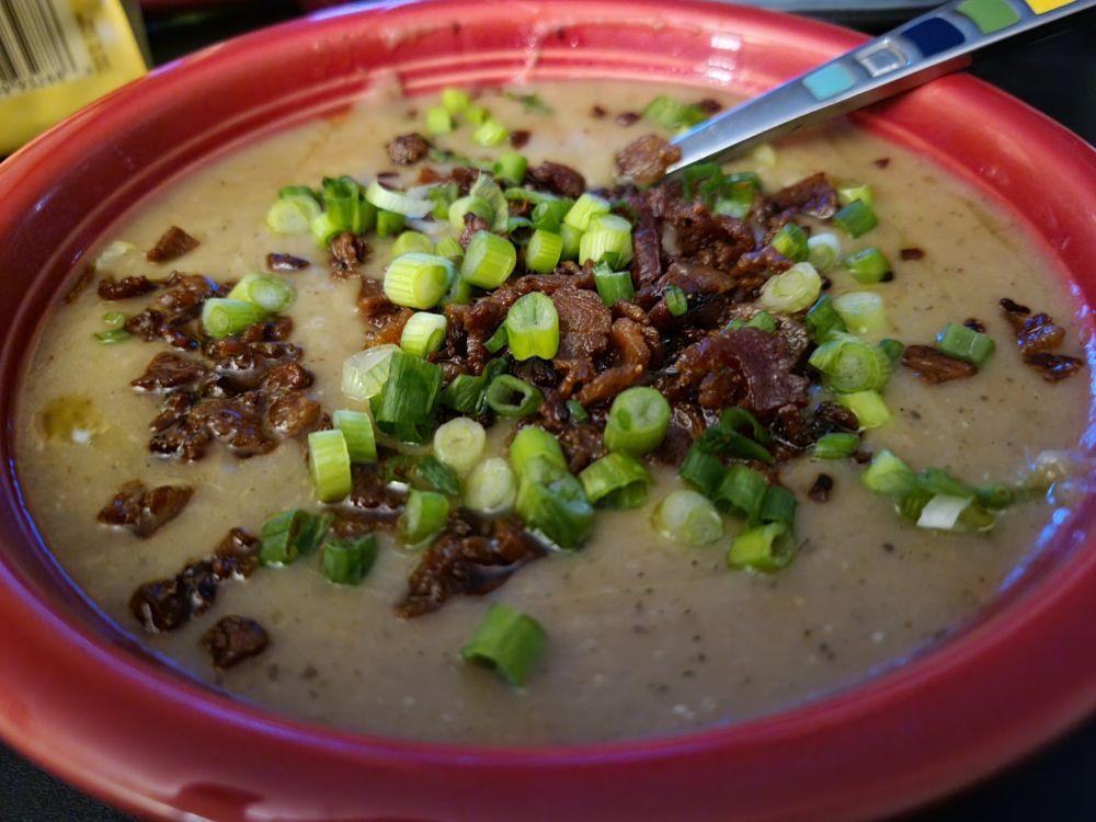 Red ceramic bowl with creamy beige soup. It is topped with small crispy pieces of bacon chopped green onions. In the back of the bowl a handle of soup soon sticks out and has colorful squares going from blue to yellow. 