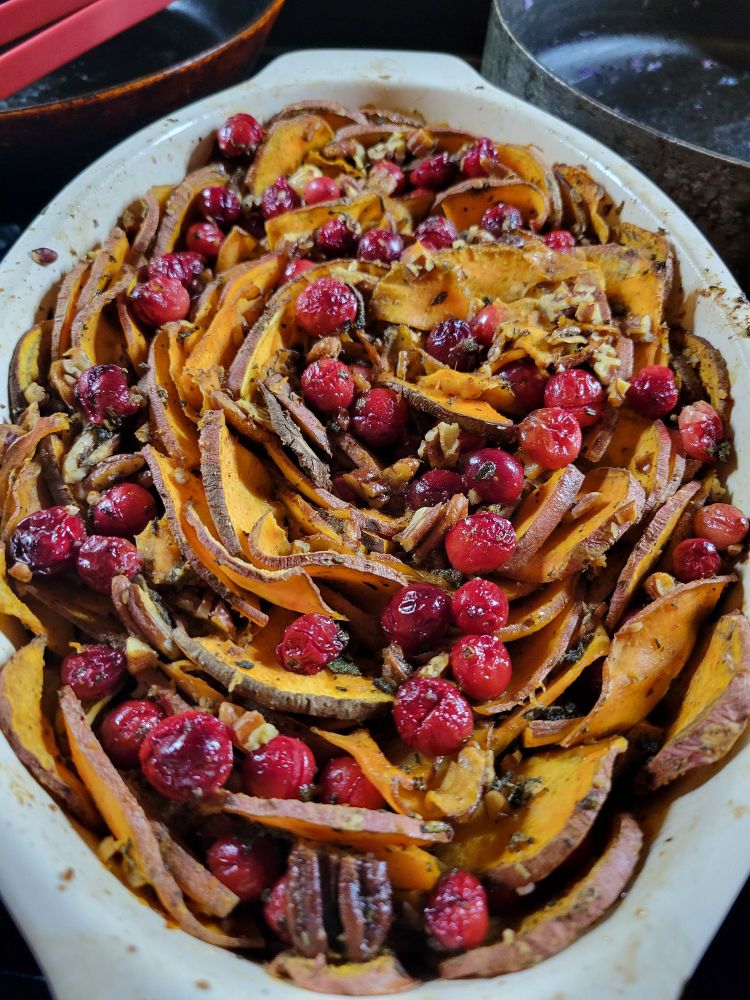 An oval dish with sliced yams arranged in a circular pattern. The yams are sprinkled with cranberries and pecans. 
