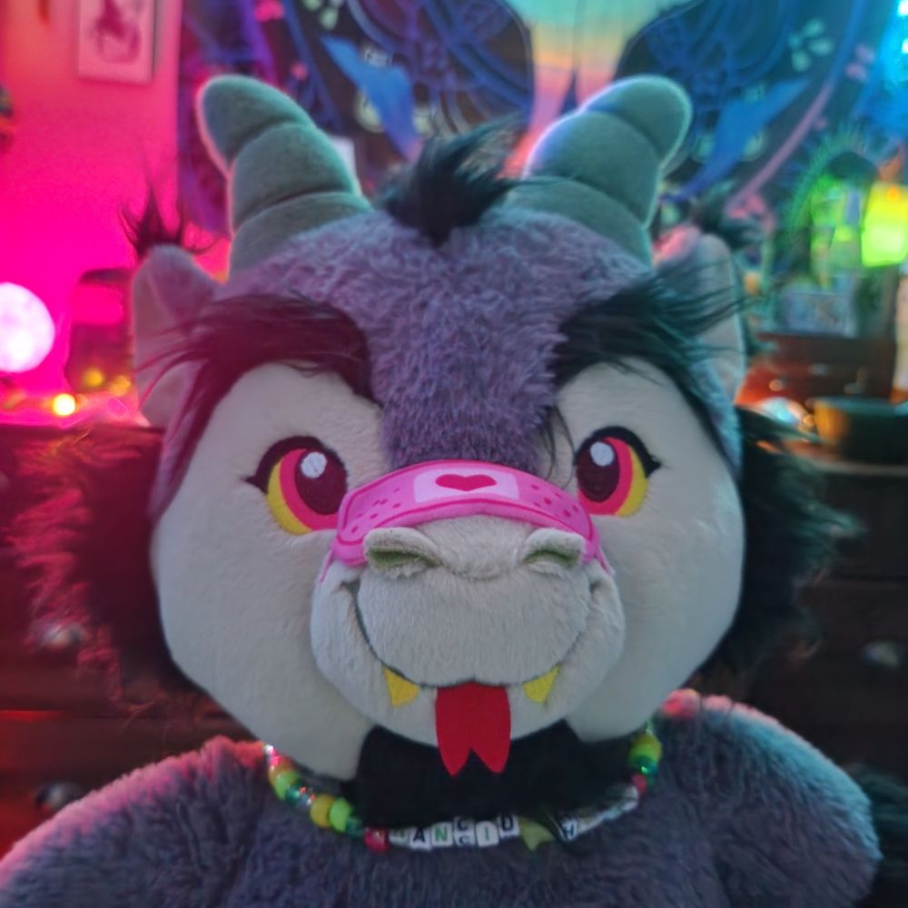 Headshot photo of a plush Rancid Heart Beelzebear wearing a kandi necklace with his name and a pink plush bandaid with a heart on his snout. Chill bedroom lighting with pinks and greens in the background 