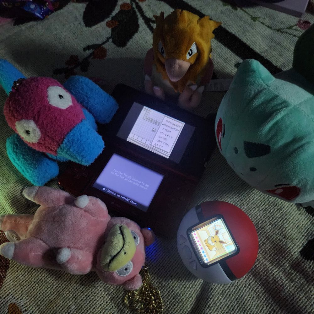 photo of a red 3ds playing Pokémon red verson, and a pokemon friends pokeball virtual pet device with an eevee on it. There are also four various Pokémon plush- porygon, slowpoke, spearow, and bulbasaur 