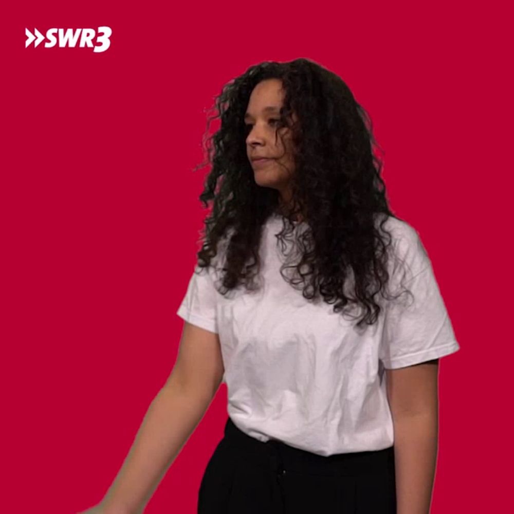 a woman with curly hair wearing a white t-shirt and black pants against a red background with swr3 written in white