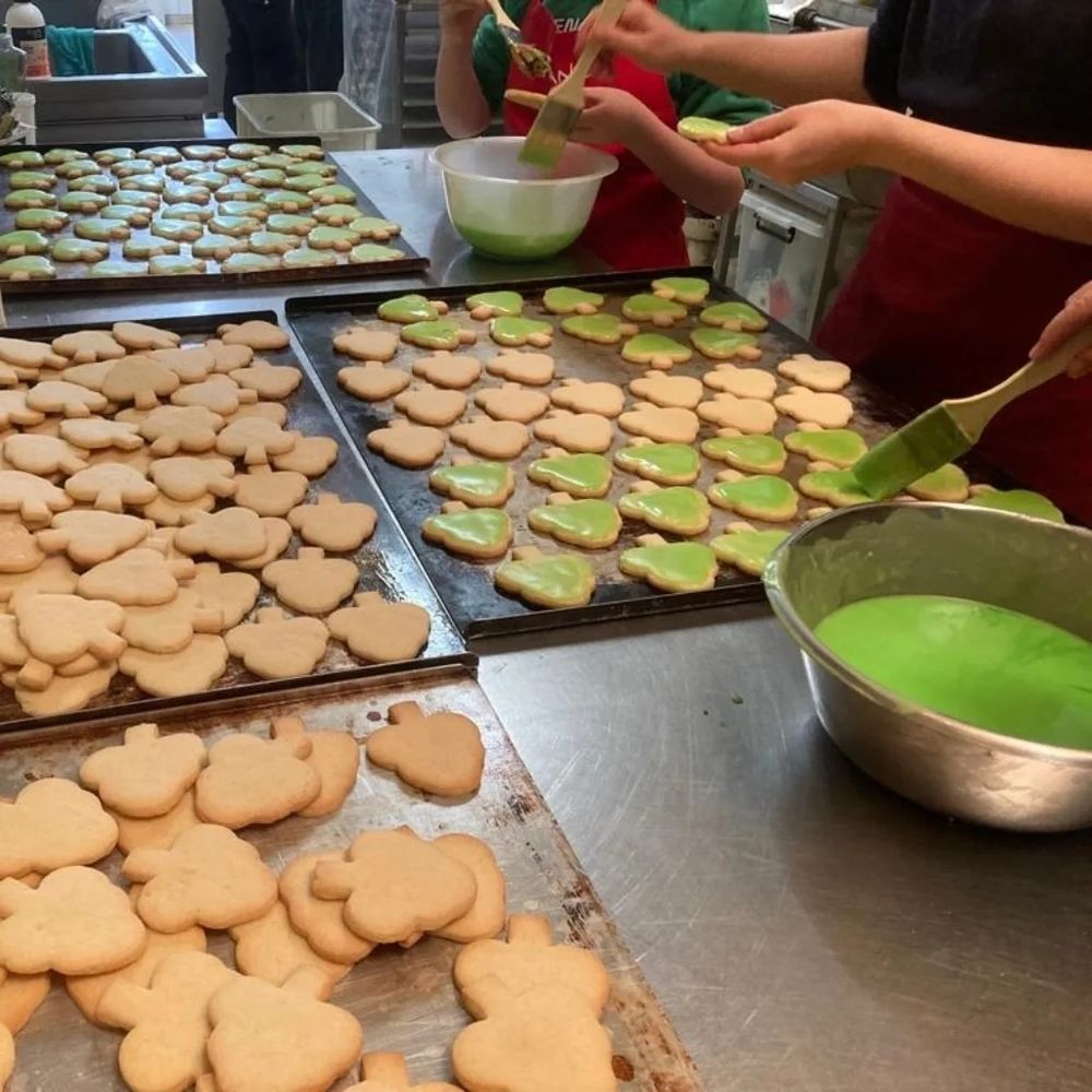 Bleche voller Gutzi (Keksen) in Baumform. Zwei Menschen sind daran, die mit grüner Glasus zu bestreichen.