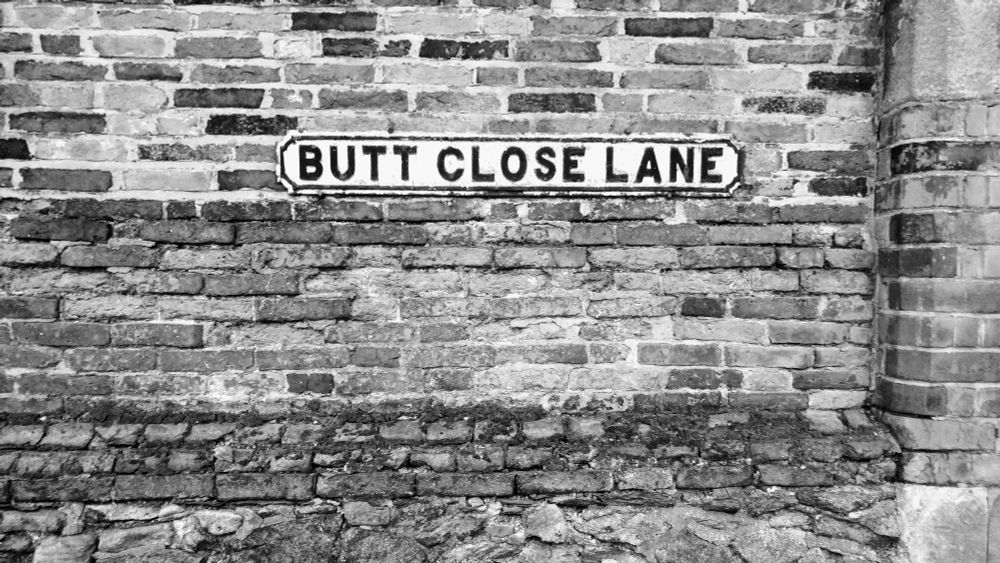 A street sign affixed to a brick wall reading "BUTT CLOSE LANE"