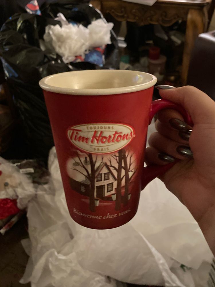 My hand holding a vintage Tim Hortons mug depicting a snowy house. The background is a living room with Christmas gift wrapping on the floor. 