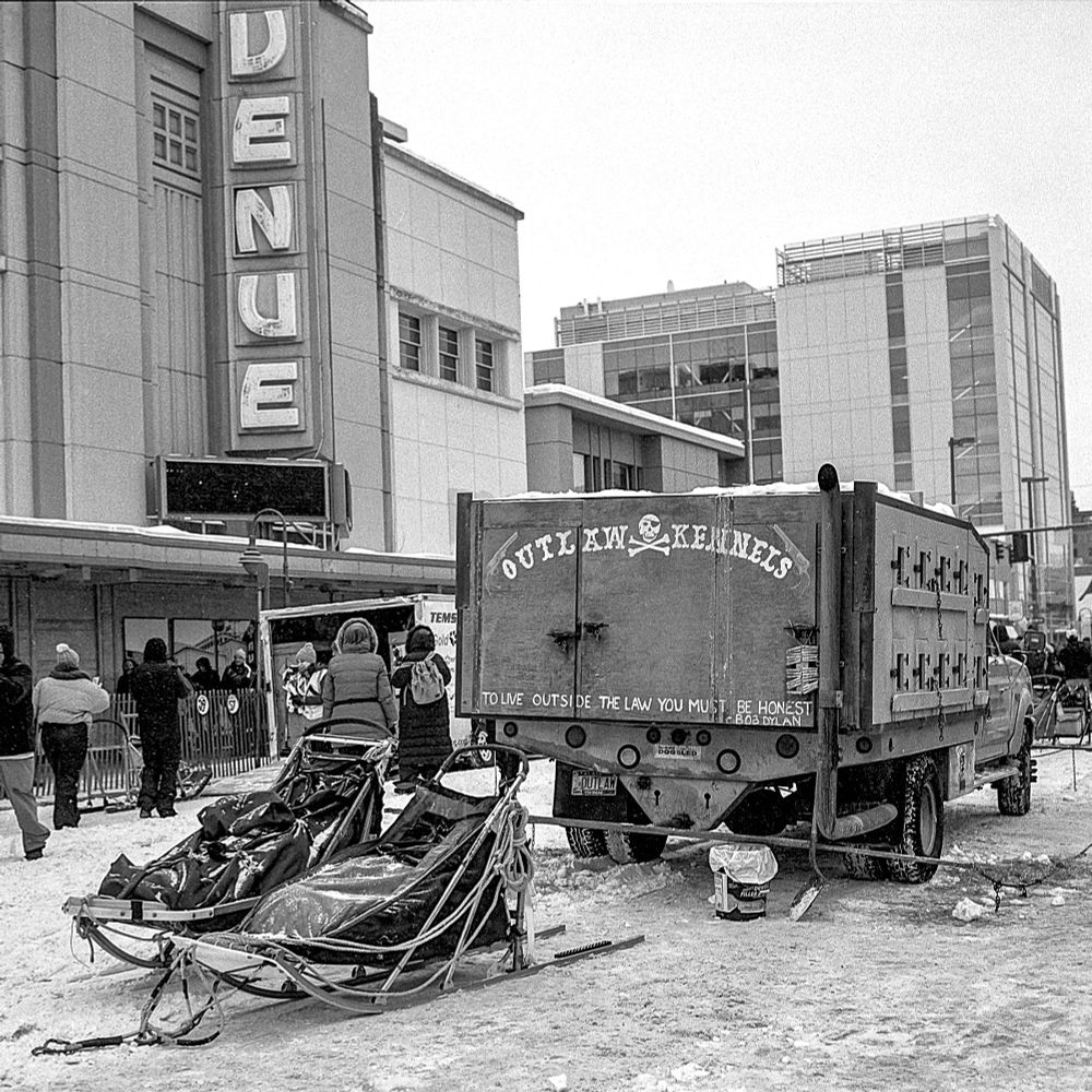 Iditarod captured on black and white film. Picture of a few sleds below the 4th Avenue sign from a few years back (they have torn the building down now... the fools). The Outlaw Kennels dog truck has an applicable Dylan quote on the back: "To Live Outside the Law, You Must Be Honest." 