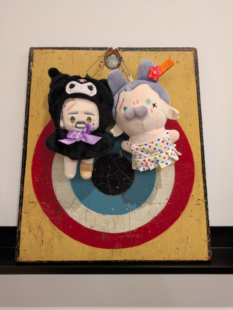 10 cm Izzy (in a Kuromi costume) and Bunny Izzy (in a handmade polka dot dress) by @imrights. They are hanging from a vintage wooden board painted with a bullseye in primary colors. 