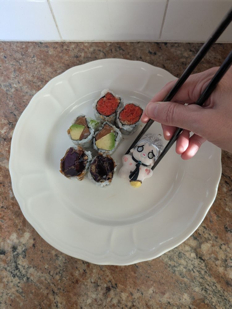 Tiny Izzy doll by @disabledpirate on a plate with six pieces of sushi. He is about to be picked up by chopsticks. 