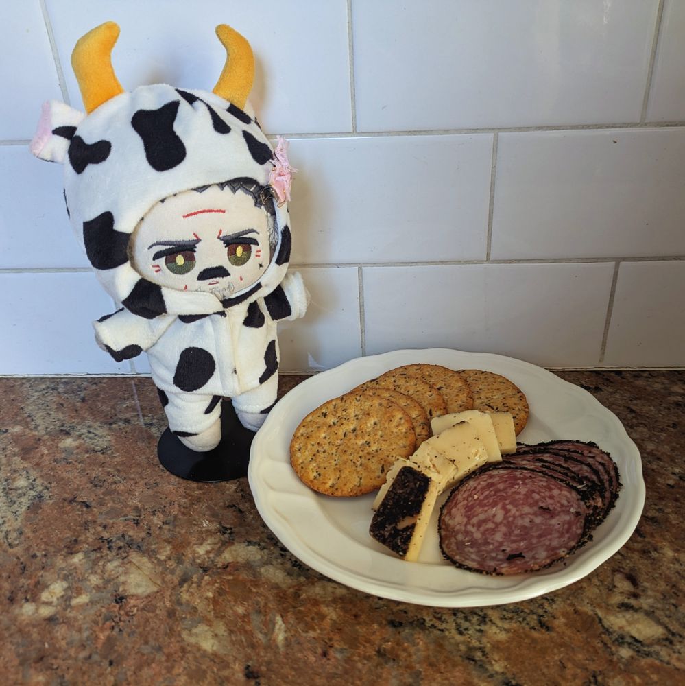 Izzy doll by @imrights wearing a cow costume. He stands next to a plate of cheese, cracker, and salami. 