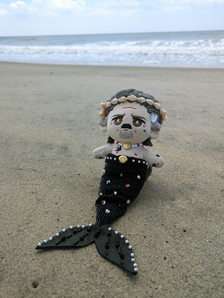 Izzy doll by @imrights wearing a mermaid outfit by @spacecadetglowao3. He has a black tail with a leather fin. The outfit is decorated with pearls, sequins, and beads. He also has a matching necklace and a shell headband/crown. He is sitting up on a sandy beach with the Atlantic Ocean in the background. 