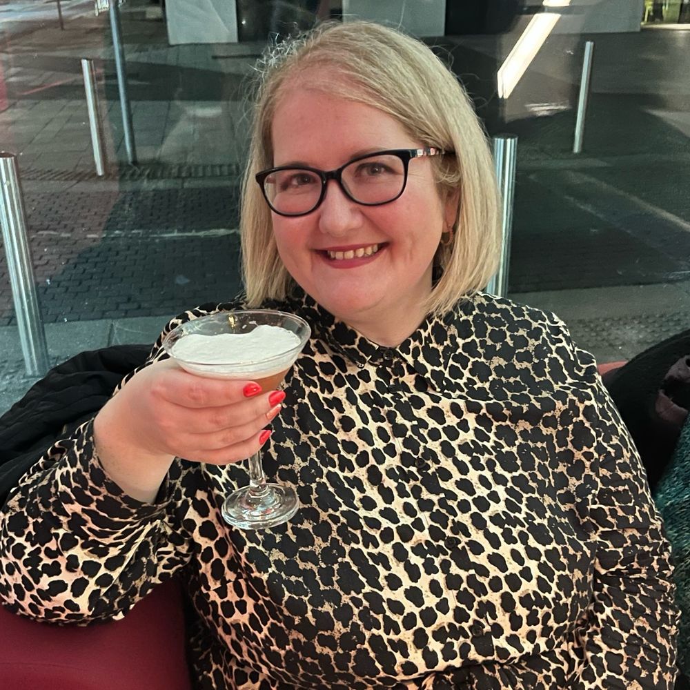 Lauren is drinking a cocktail from a martini glass. She has short blonde hair and wears thick dark-rimmed glasses. She is wearing a high-necked, long sleeved leopard print dress. She is sitting in the foyer of a theatre. She is smiling widely. 