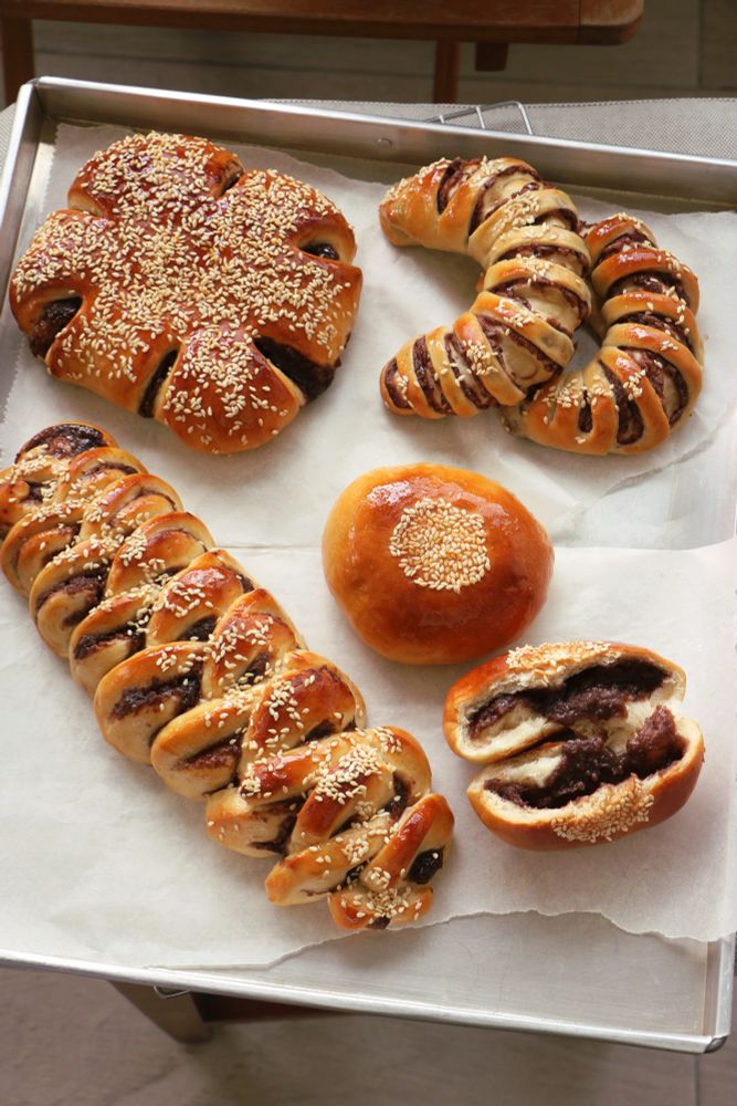 four kinds of red bean buns on a tray: one flower-shaped, two striped crescents, two buns with one split open to show red bean paste, and one braid made of alternating cinnamon-roll-like rounds of dough and bean