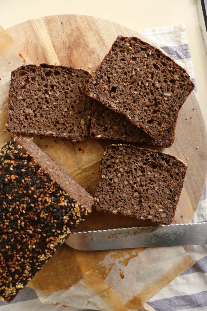 a round wooden board with a loaf of sesame-roofed bread, with several dark, square slices with a webby crumb and a seeded, nutty, grain-pocked complexion, and a serrated knife