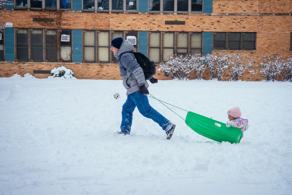A father dragging his kind on a sled by a school, both enjoying themselves