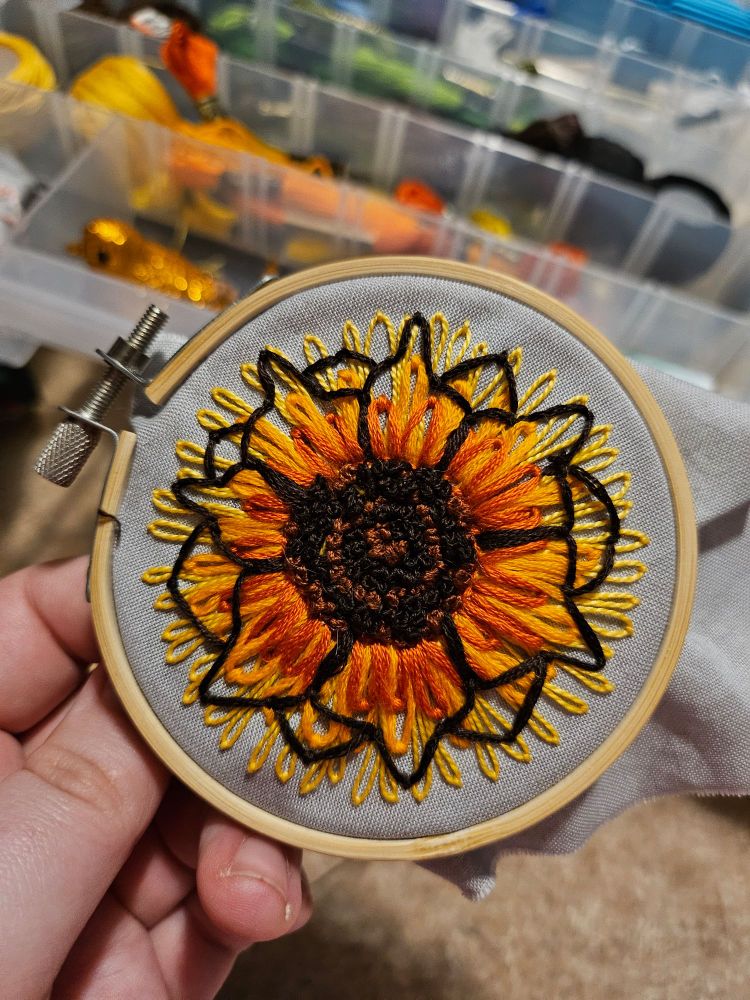 palm-sized embroidered sunflower (little smaller than the other one). the petals are shades of orange near the center and transition to yellow at the tips. the middle of the flower is brown and black(maybe a darker brown actually? i dont remember) in alternating rings.