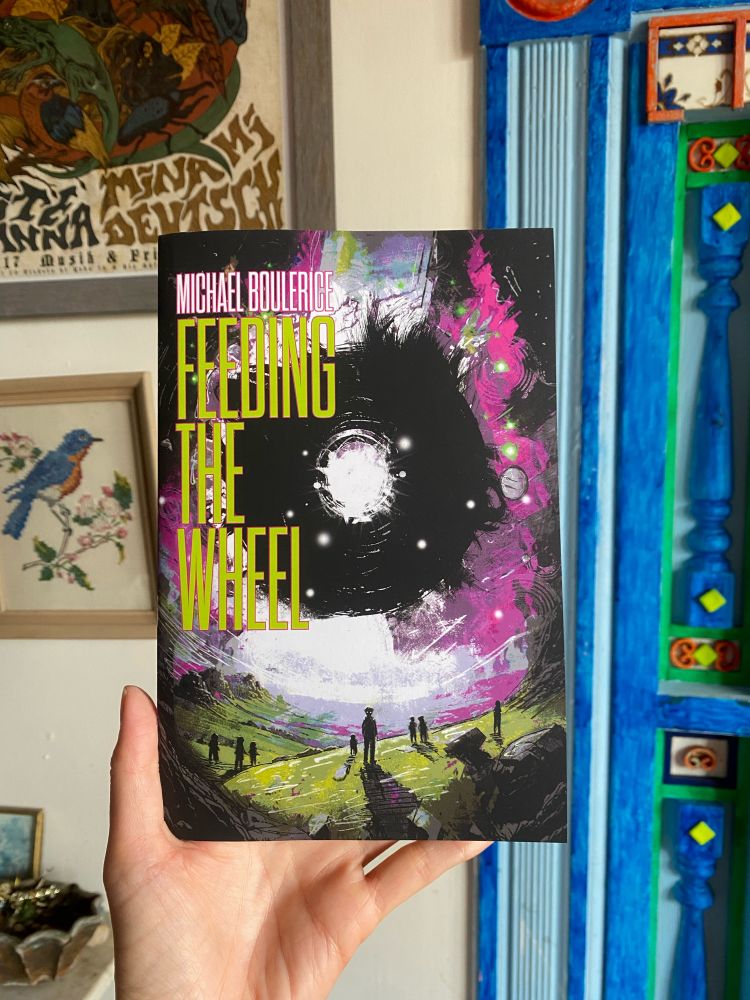 Hand holding up the novella Feeding the Wheel by Michael Boulerice. The cover features children standing on a green rocky hill in front of a night sky with a white circular hole or portal in it.