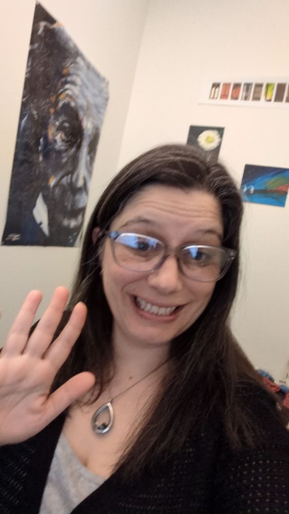 A woman with glasses and long brown hair waving hi and smiling. I'm the background is a poster of Albert Einstein and some paintings of flowers and sailboats. 