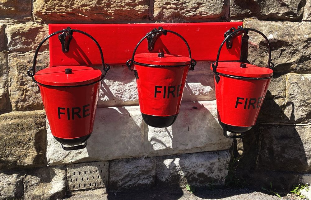 Three red fire buckets with the word fire on each on a red board against a stone wall. 