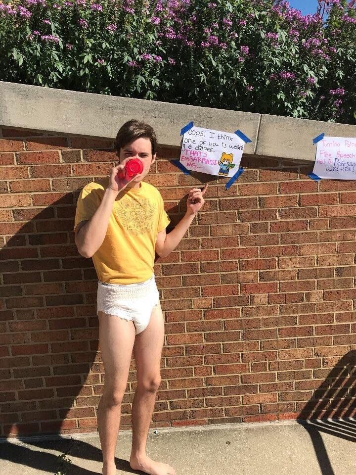 Photo of Charlie Kirk in a diaper drinking from a bottle during a college protest. He's pointing to a sign that reads "Oops! I think one of you is wearing a diaper. That's embarrassing" 