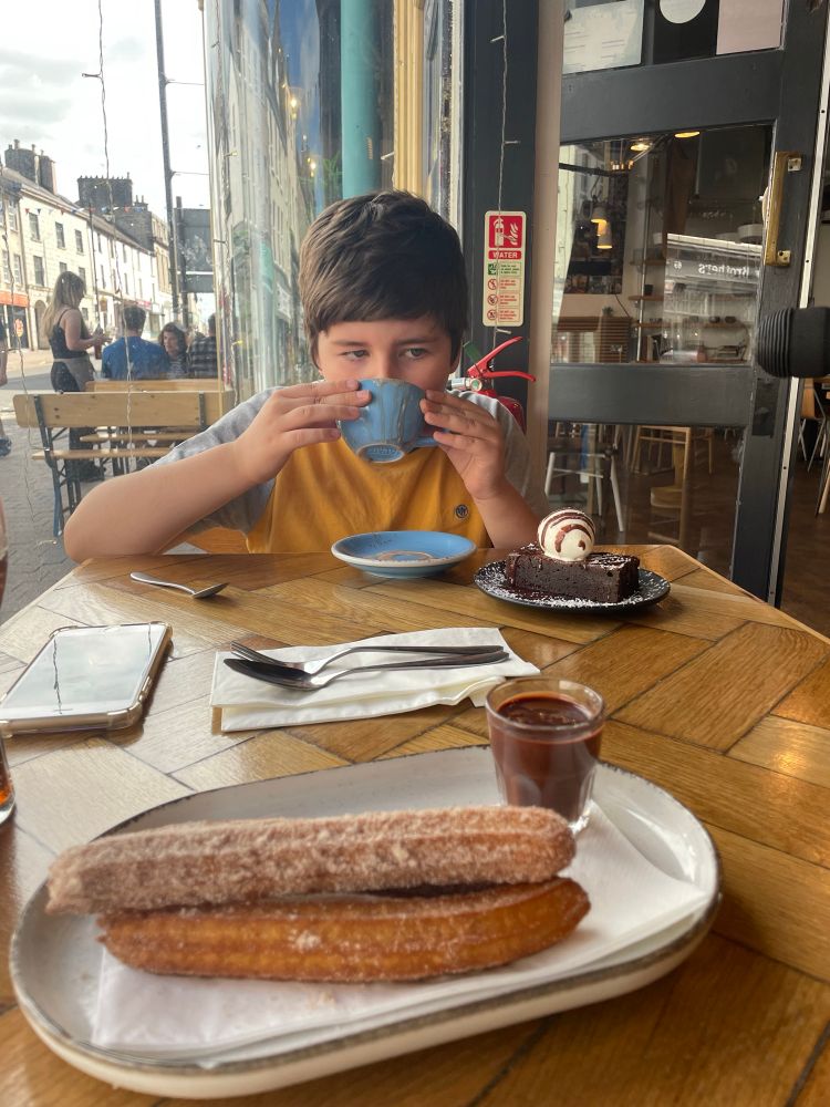 Little boy drinking a blue mug of hot chocolate with a chocolate brownie arms a plate of churros on the table