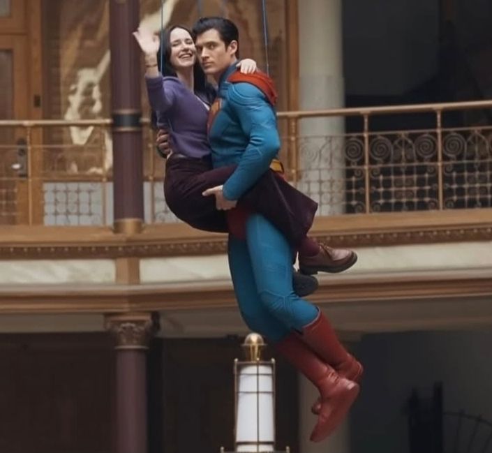Ator David Corenswet caracterizado como Superman, segurado no ar por cabos, segurando a atriz Rachel Brosnahan, caracterizada como Lois Lane, usando calça comprida marrom escuro puxado pro vinho, sapatos marrom mais claro, e blusa roxa. Rachel está no colo de David, que está a segurando com uma mão na coxa e outra nas costas. Ambos estão suspensos no ar por cabos.