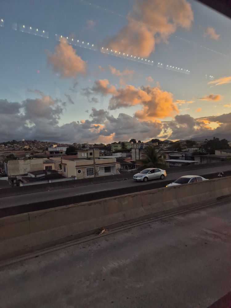 Foto mostrando uma avenida com carros passando, mas o foco está no céu ao fundo, azul de fim de tarde com algumas nuvens branquinhas na luz meio alaranjada