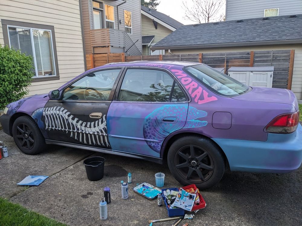 A mostly purple car with hand painted panels of blue and black and illustrations on it. Art supplies are on the foreground.