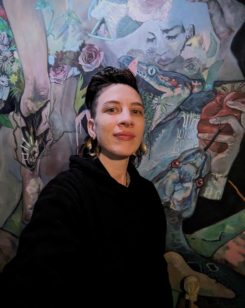 A photo of me in a black hoodie in front of my latest work in progress, a large oil painting. Two snakes on the right side along with a detached hand holding a heart on the right. On the left is a figure, partially disjointed, its forearm is breaking open to reveal roots and mushrooms growing within.