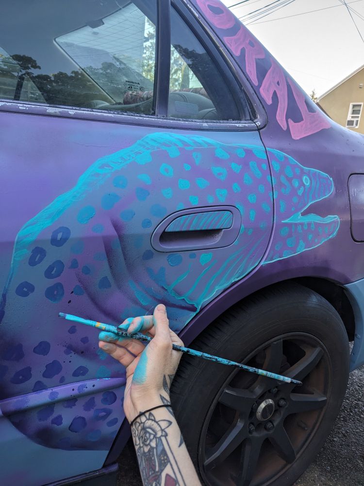 A hand painted teal eel on the side of a purple car. A hand with teal paint on it is holding a paint brush up to the car/eel.
