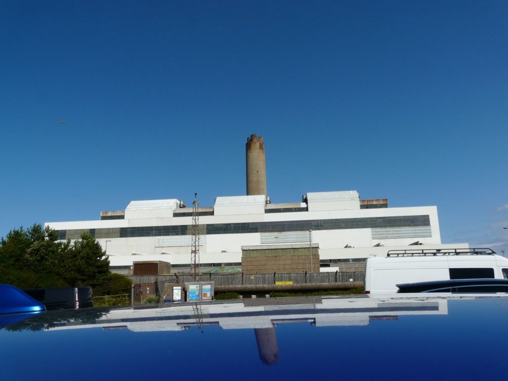 A disused power station against a blue sky, also reflected in the roof of a blue car