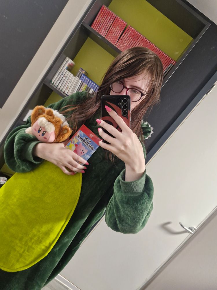 A selfie of me in a Dino onesie holding Kirby Air Riders and Animal Kirby plush.