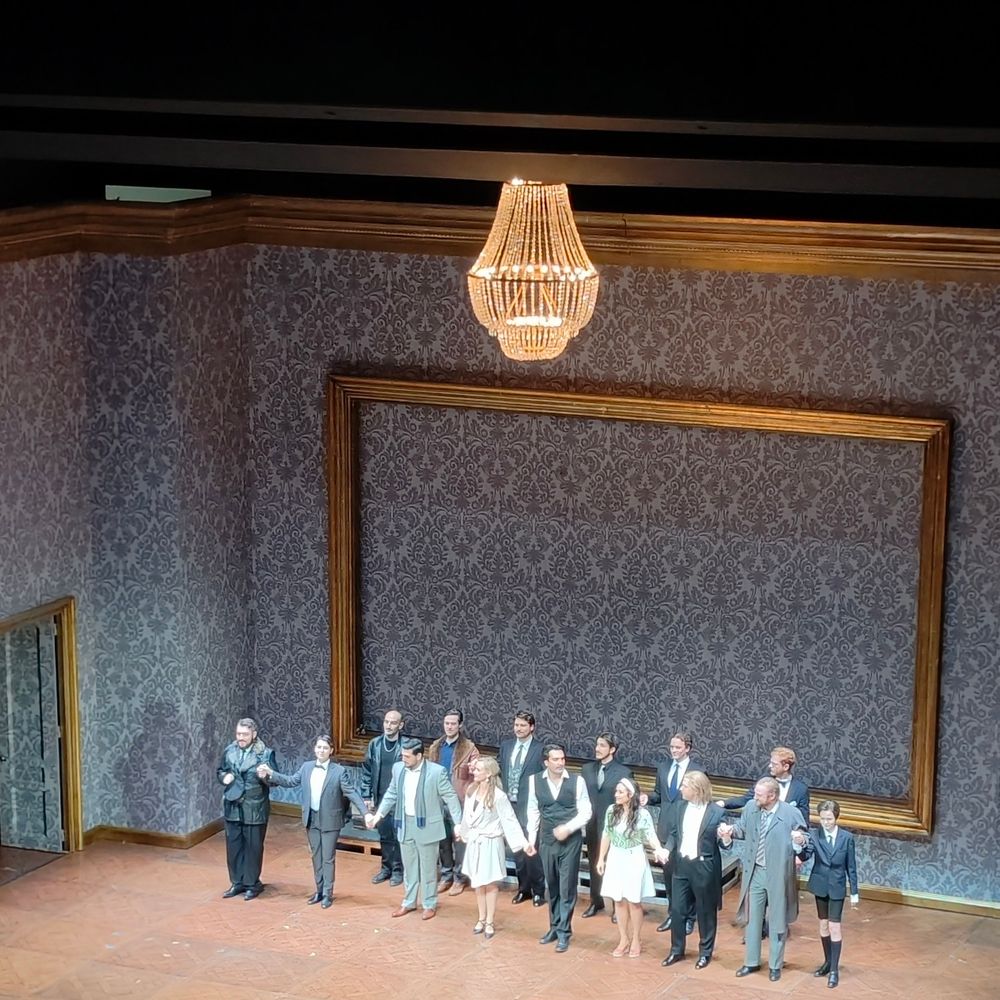 Eine Gruppe von etwa 15 Schauspielern in Abendgarderobe hält Händchen und verbeugt sich auf einer Bühne nach einer Aufführung. Im Hintergrund ist ein großer, leerer Goldrahmen an einer grauen, gemusterten Wand zu sehen, beleuchtet von einem prunkvollen Kronleuchter.

