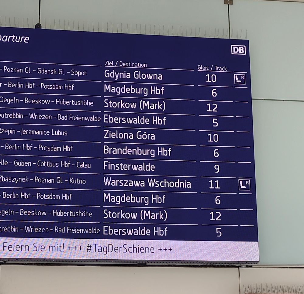 Blaue Abfahrtstafel eines Bahnhofs. Vor heller Wand mit deutschen und polnischen Ortsnamen.