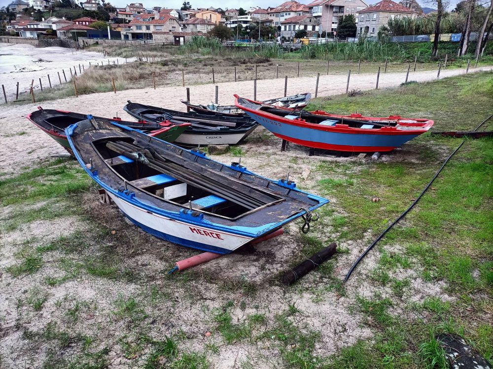 Unhas barcas nunha praia en Coruxo. A primeira é branca e azul e ten escrito o nome " Merce" despois hai outra negra e branca co nome " Chisca " e máis lonxe unha vermella e azul.