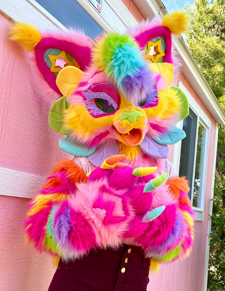 A bright pink and rainbow cat fursuit with glittery pink eyelashes and rainbow flower petals around her face. His paws have rainbow claws and heart shaped stripes. The entire fursuit is covered in dark pink and rainbow stripes. The ears have star shaped gauges. 