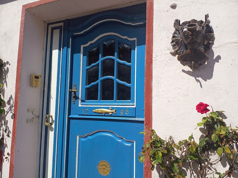 Blaue Tür mit Sprossenfenster im oberen Bereich mit messingfarbenen Verzierungen und einer Messingtürklinke. Das Haus ist weiß gestrichen, die Türöffnung rot umrandet. 