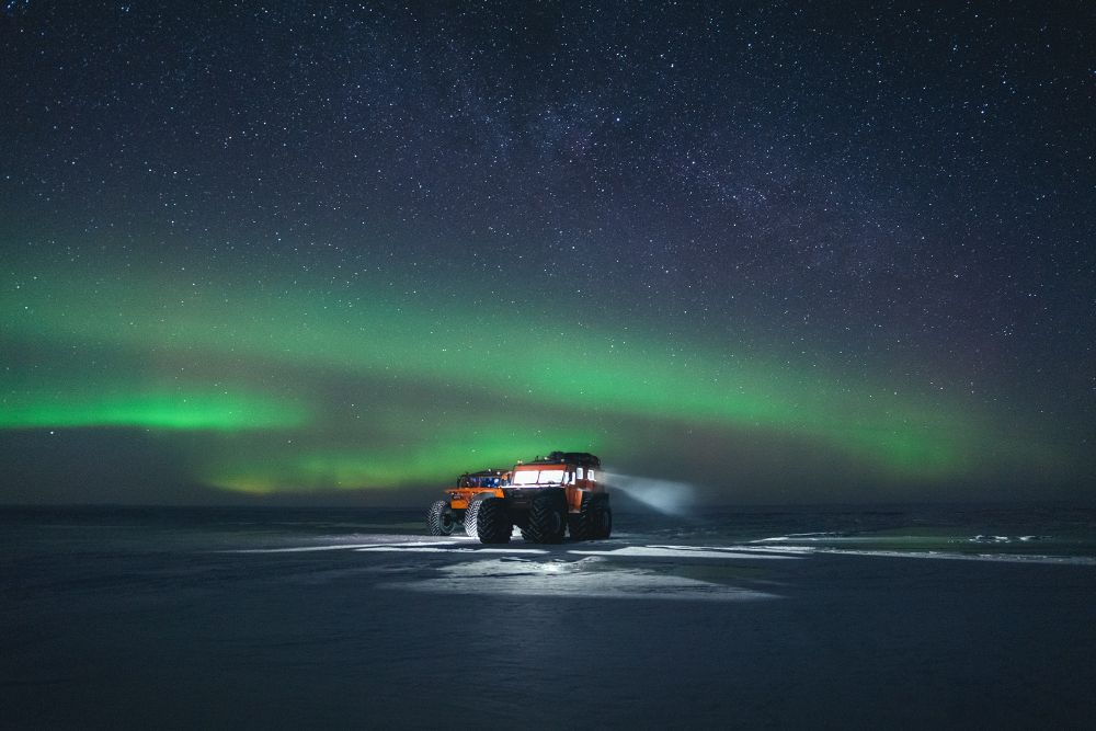 Photograph. It appears to be night. A vast plain of flat, tightly packed ice and snow extends to to the horizon. A green aurora stripes across the sky in three distinct bands. Beyond the aurora the sky is full of stars. A truck, large and yet viewed from such a distance looks small and soinsignificant. The lights of the truck are ablaze with thick spotlights shining out onto the ice, creating a small pool of light around it.
