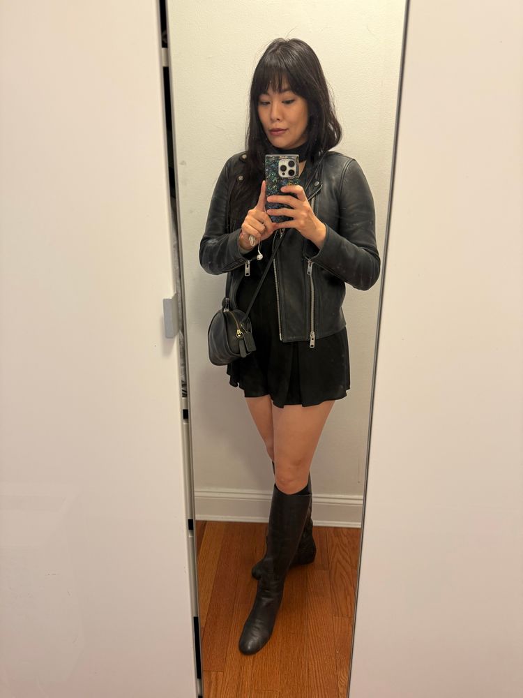 Mirror selfie of a girl wearing a leather jacket, small crossbody purse, short romper, and knee high boots, all in black