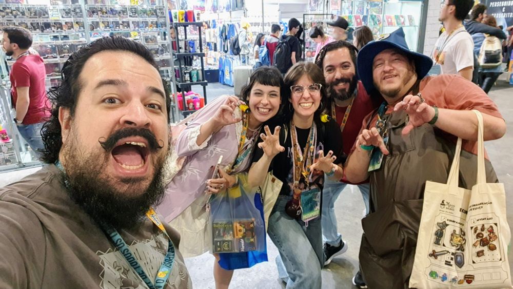 A imagem é um selfie tirado de um grupo de cinco pessoas, todas sorrindo e olhando para a câmera, em um ambiente que parece ser uma feira ou evento movimentado com muitos expositores ao fundo.

Edu: Em primeiro plano, no canto inferior esquerdo, está o homem com barba e bigode pretos longos e cabelos escuros, que aparece em outras fotos. Ele está usando uma camiseta cinza e tem um cordão de crachá no pescoço. Sua boca está bem aberta, expressando um grito de alegria ou surpresa.

Bianca: Ao lado dele, no centro, está uma mulher jovem com cabelos curtos e escuros. Ela está sorrindo, vestindo um casaco lilás sobre uma blusa clara e segurando uma sacola azul e transparente com um jogo de tabuleiro dentro. No pescoço, tem um crachá de evento.

Marjô Rui: À direita dela, outra mulher jovem, com cabelos castanhos e óculos de armação clara, também sorri amplamente. Ela faz um gesto com as mãos, com os dedos curvados como garras. Ela veste uma camiseta escura e jeans, e também tem vários cordões de crachá no pescoço.

Lucas Gandra: Atrás das duas mulheres, e entre o homem de barba e o homem do chapéu, está um homem com barba e cabelos escuros, sorrindo gentilmente. Ele veste uma camiseta vermelha e tem um cordão de crachá.

Blonde: No canto direito da imagem, está um homem usando um chapéu azul pontudo, como um chapéu de mago, e uma camiseta coral. Ele está fazendo um gesto com as mãos, como se estivesse tentando assustar, com as mãos abertas e os dedos curvados. Ele tem um avental ou macacão escuro e carrega uma sacola de tecido claro com uma ilustração na lateral.

O fundo da imagem está repleto de stands, prateleiras com produtos como caixas de jogos ou colecionáveis, e outras pessoas circulando, indicando um ambiente de convenção ou feira. A iluminação é clara e uniforme.