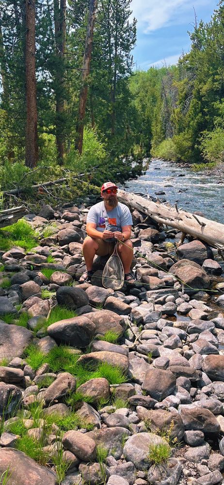 A picture of me with a smile on my face, tying a fly to a fishing line. I’m sitting on a rock next to a flowing mountain river. 