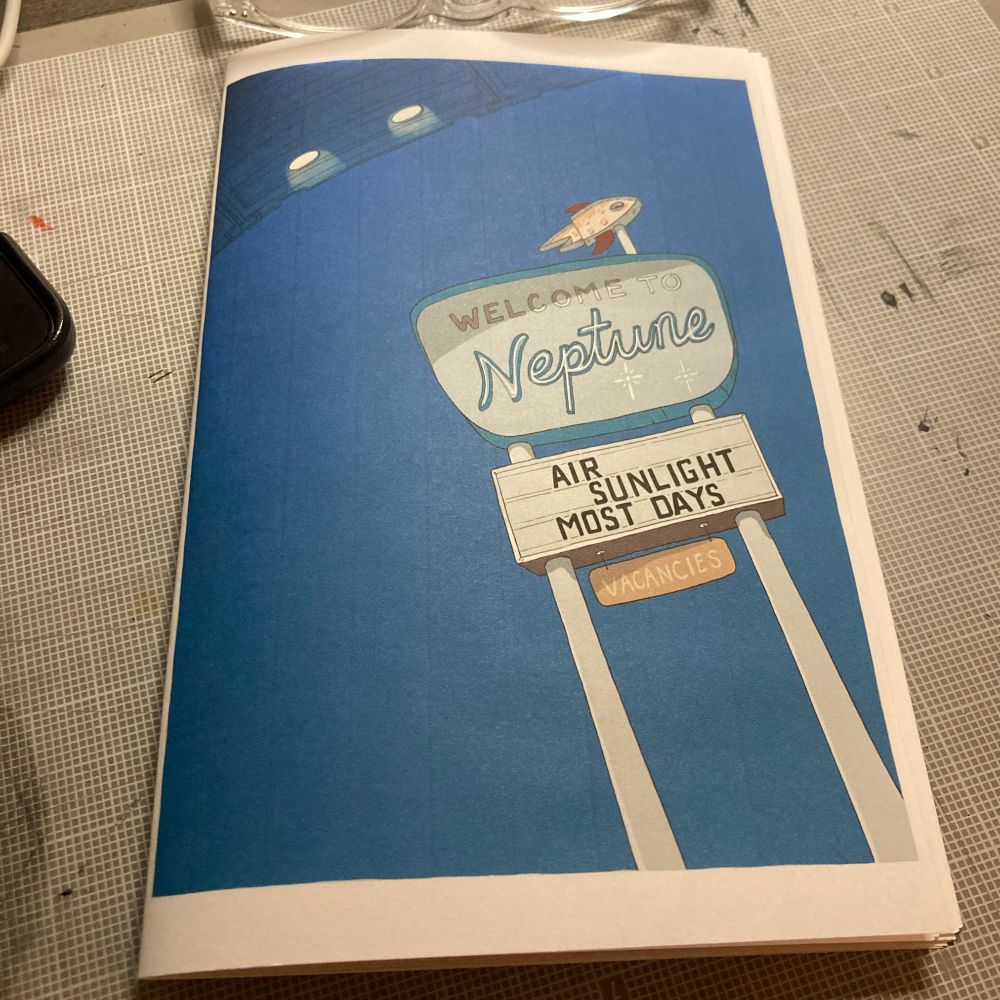 Photo of the front cover of Neptune Road, depicting a roadside sign that reads "Welcome to Neptune, air sunlight most days, vacancies" and some sort of transport flying overhead