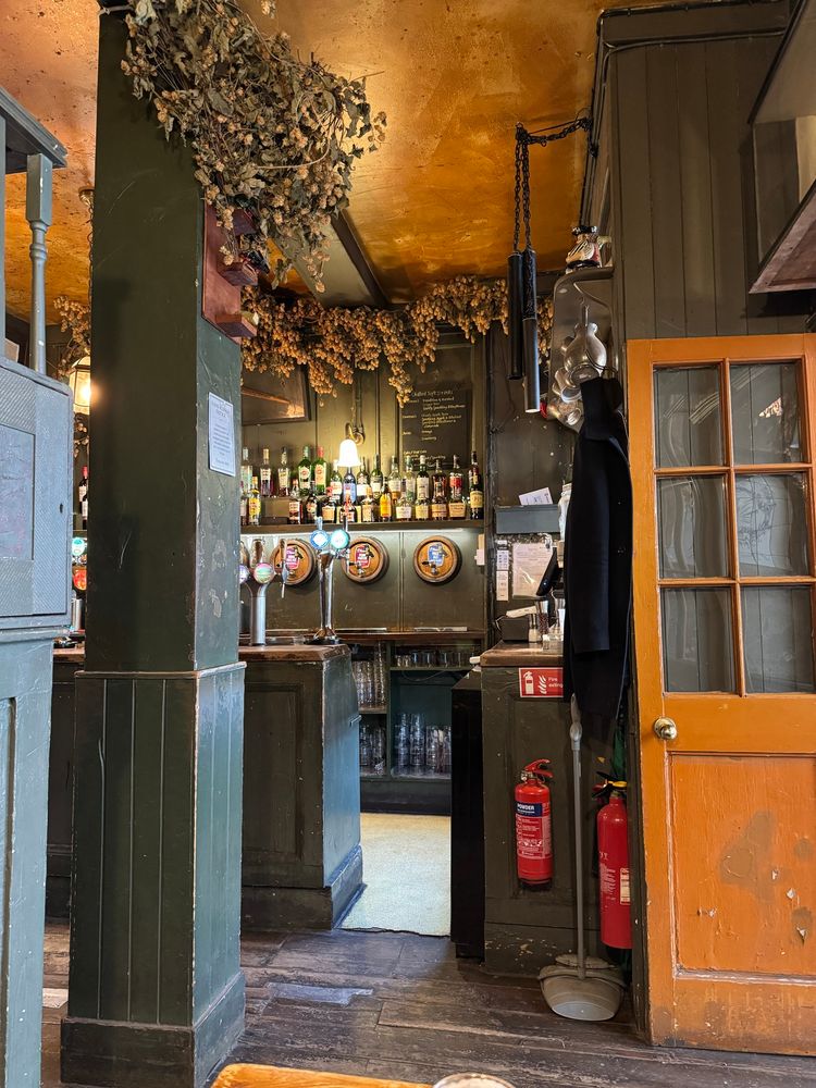 A cozy pub interior with dark green wooden paneling, an orange door, and a wooden floor. The bar area features several bottles of liquor on shelves, beer taps, and glassware. Dried hop vines hang from the ceiling, and there are fire extinguishers and a coat hanging near the entrance. The atmosphere is warm and rustic.