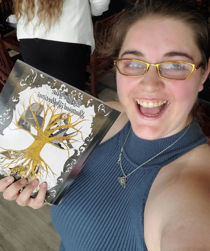 Jerrica with an open mouthed smile holding up a copy of Shattered Reflections: By the Fire.