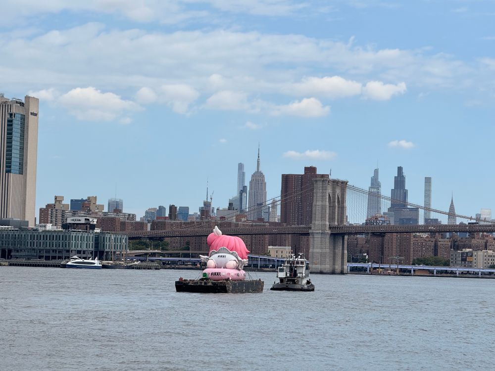 The east river. In the foreground there’s a pink inflatable bull (maybe?) on top of another white animal on a pink inflated raft all on a small barge, the background is the Brooklyn Bridge and Empire State Building and Manhattan skyline
